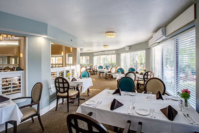 Bright dining room with neatly set tables covered in white linens, chairs, large windows with blinds, and soft ceiling lights.
