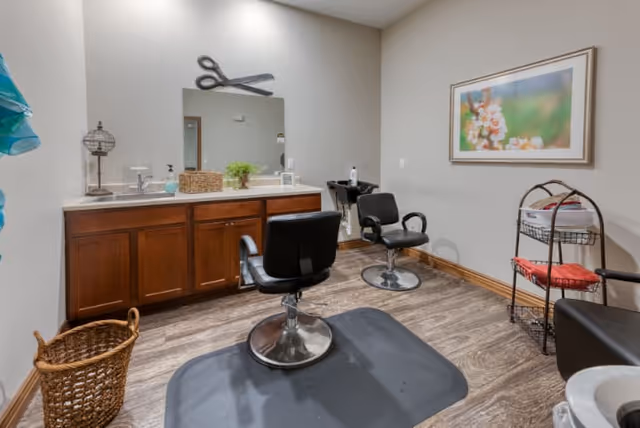 Interior view of a hair salon area with two black salon chairs on mats, a wooden cabinet with a countertop and a mirror above it, a wicker basket on the floor, a small metal rack with towels and supplies, and a framed floral picture on the wall.