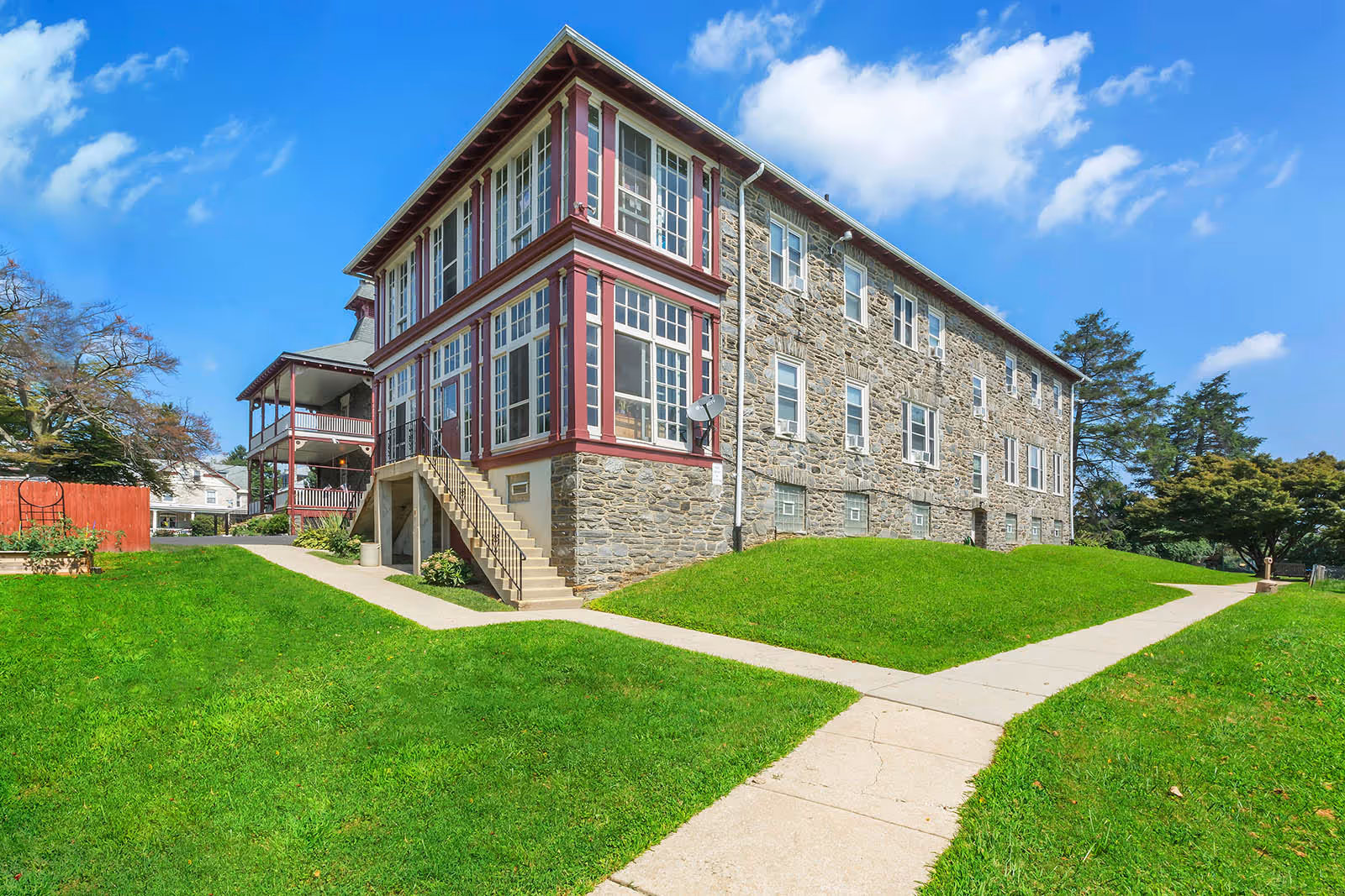 Exterior view of a large stone building with multiple windows and a red and white porch area. The building is surrounded by well-maintained green grass and concrete walkways under a bright blue sky with some clouds.