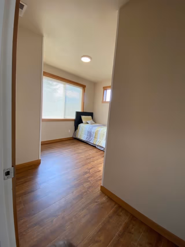 View through a doorway into a bedroom with wooden flooring, a single bed with a black headboard, and a window with blinds letting in natural light.