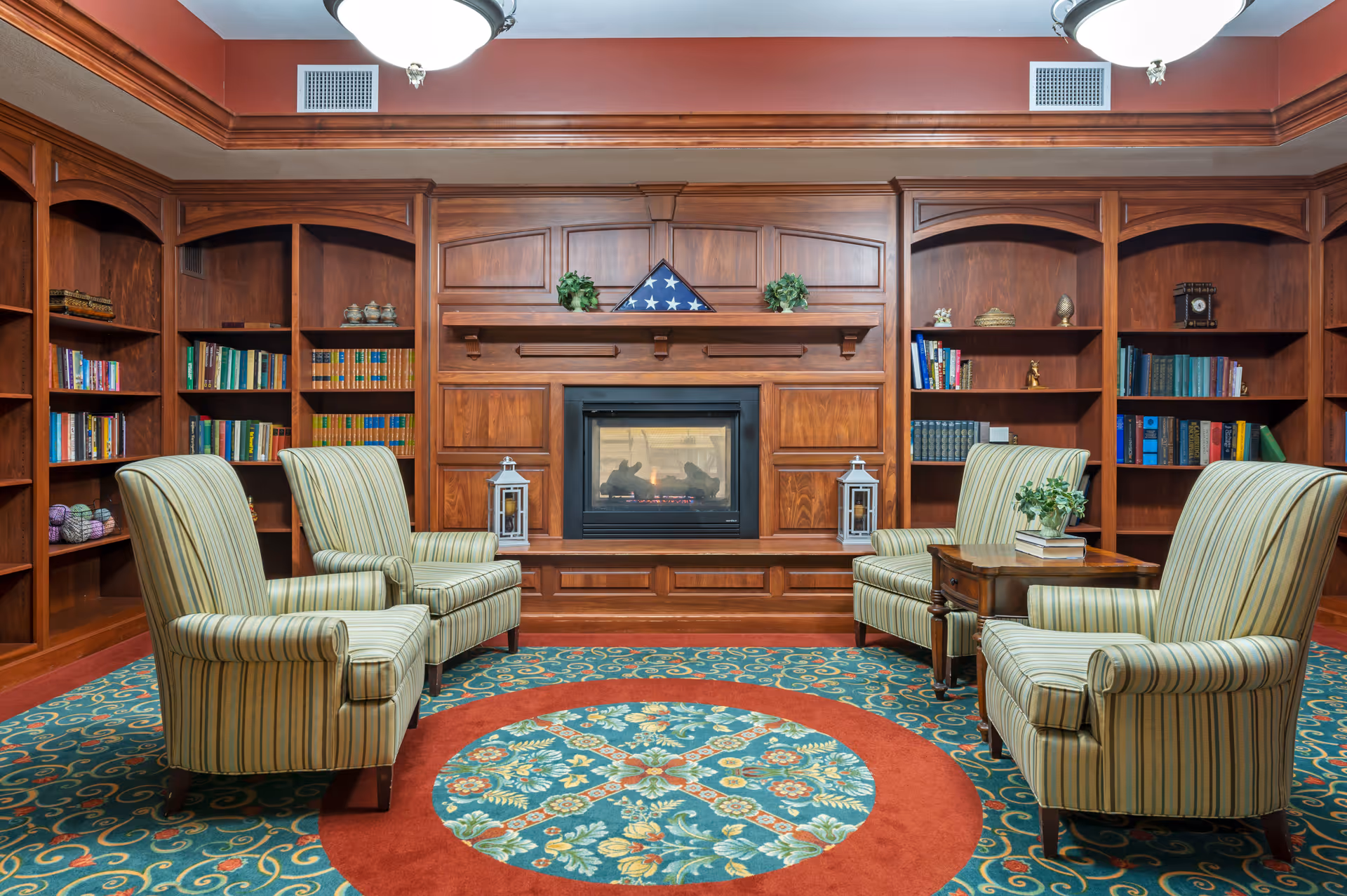 A cozy library or sitting room with four striped armchairs arranged around a wooden coffee table on a colorful patterned carpet. The room features built-in wooden bookshelves filled with books and decorative items, and a central fireplace with a folded American flag and two lanterns on the mantel. The ceiling has crown molding and two ceiling lights.