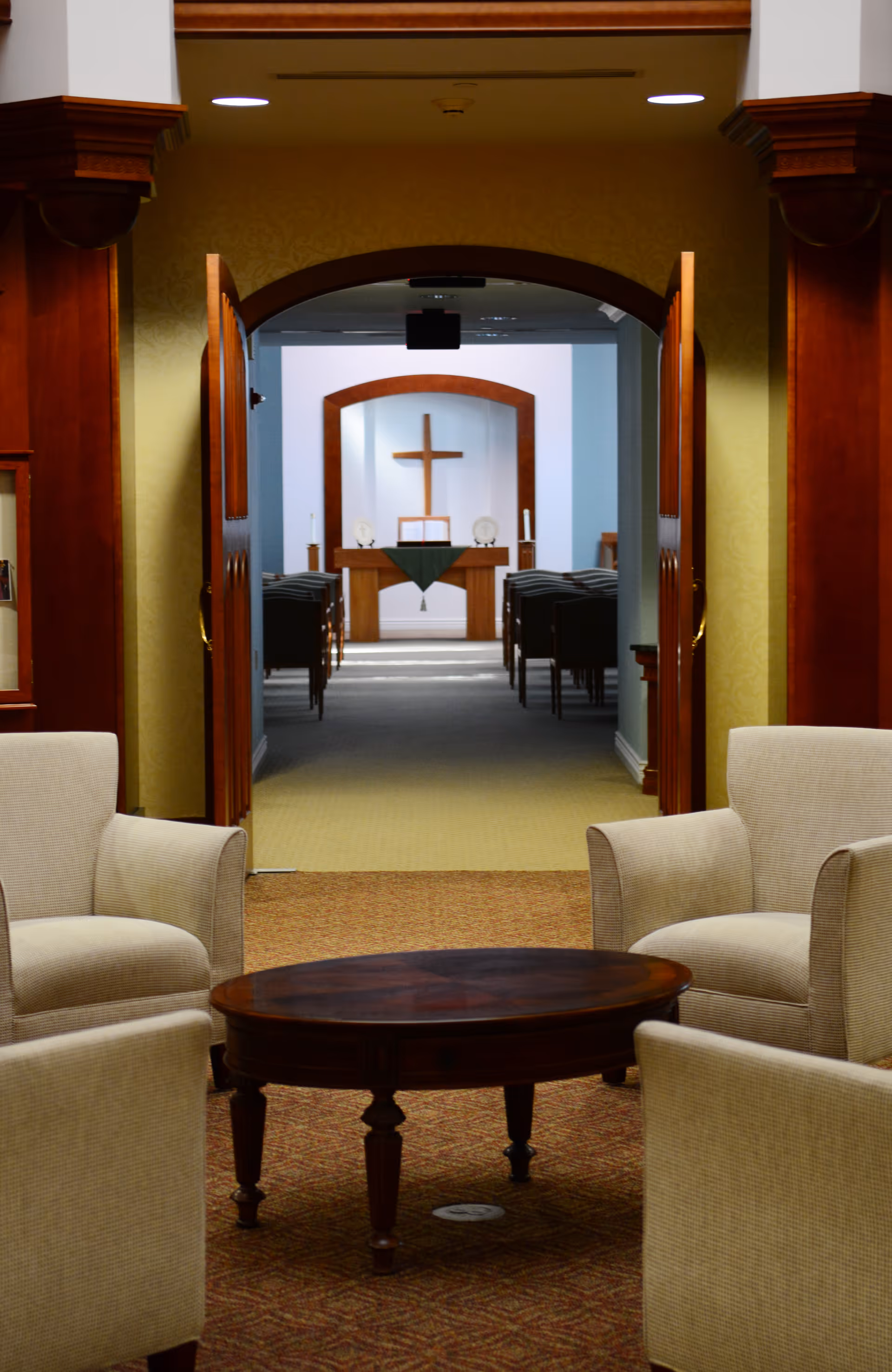 A seating area with armchairs and a round table looking through open double doors into a chapel with an altar and cross.