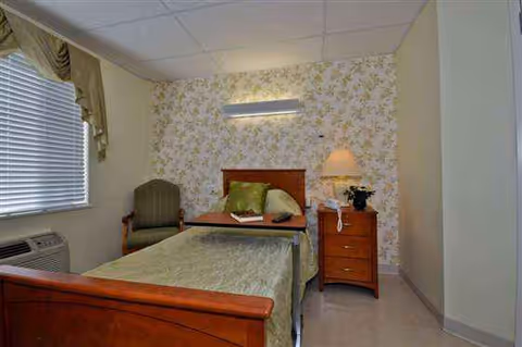 Bedroom with a single bed, dresser, chair, lamp, and floral wallpaper.