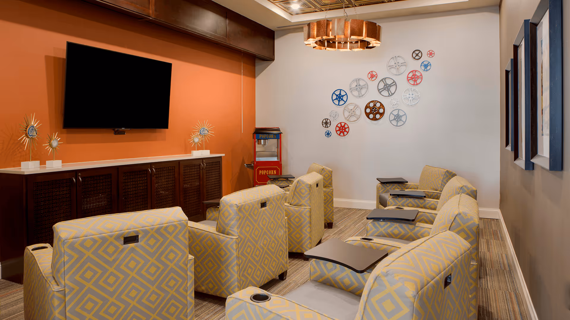 A cozy media room with six yellow and gray patterned armchairs arranged in two rows facing a wall-mounted flat screen TV. The room has an orange accent wall behind a dark wooden cabinet, a popcorn machine in the corner, and decorative film reel wall art on a white wall. A modern circular chandelier hangs from the ceiling.