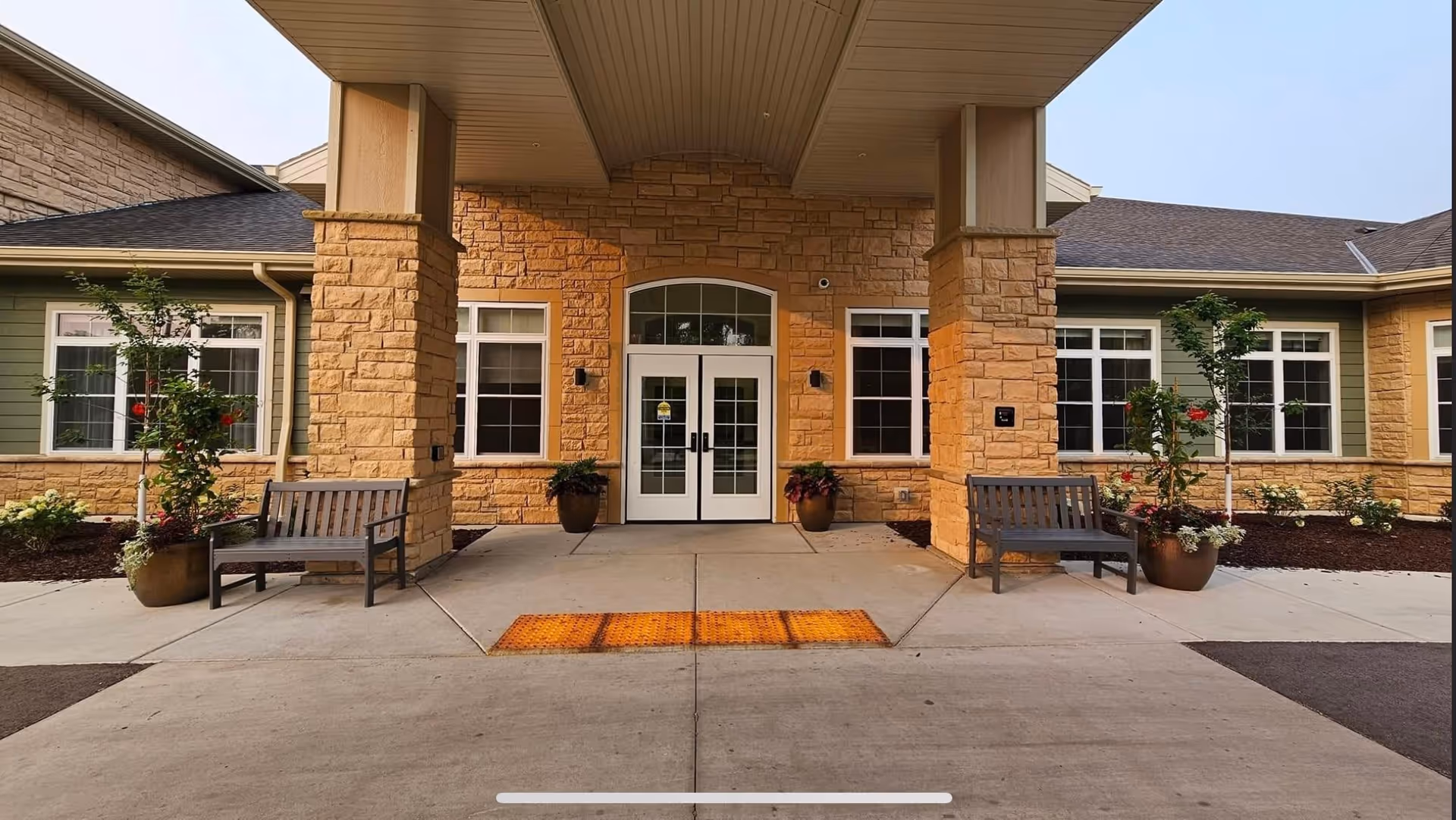 Entrance of The Courtyard at Sussex facility showing a covered walkway with stone pillars, two benches on either side, potted plants, and double glass doors with windows on both sides.