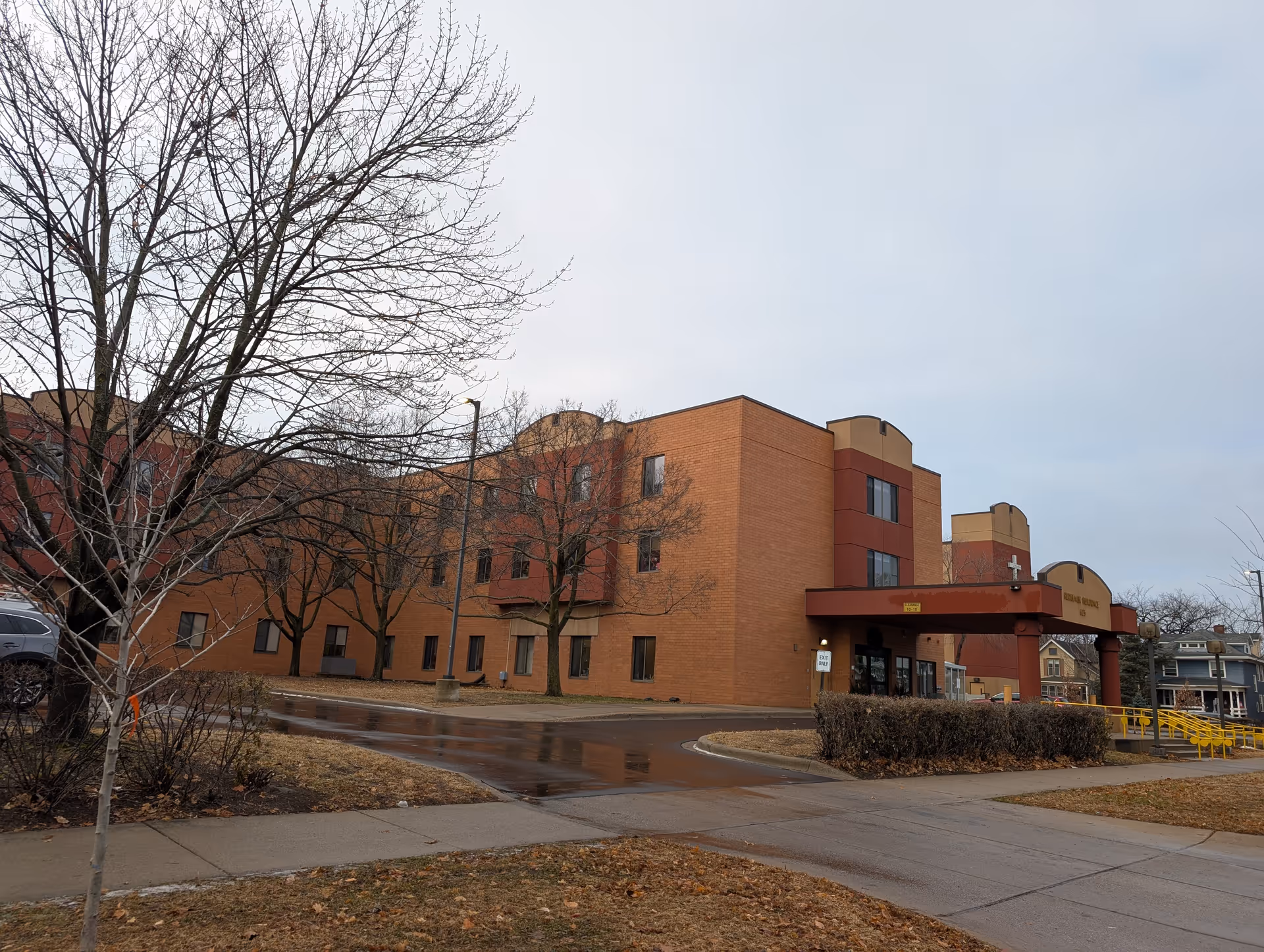 Exterior view of a three-story brick building with a covered entrance featuring a cross symbol. Leafless trees and a wet driveway are visible in front of the building under an overcast sky.