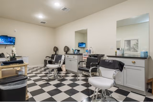 Interior of a senior living facility hair salon with black and white checkered floor, three salon chairs, two hair dryer chairs, a large mirror, cabinets with hair care products, and a small wall-mounted TV.