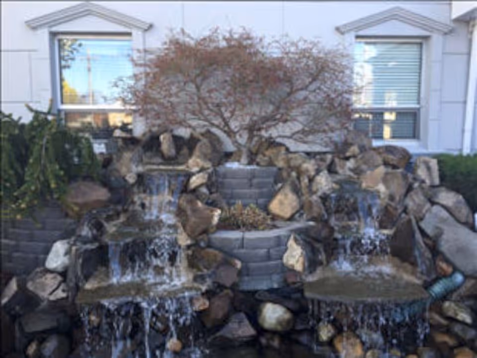 Outdoor water feature with cascading waterfalls made of rocks and stone tiers, surrounded by plants and a small tree, in front of a building with two windows.