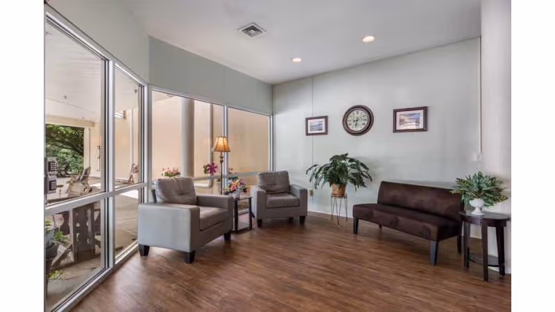 A bright and clean sitting area with two gray armchairs and a dark brown sofa arranged around small side tables with flowers. The room has large windows letting in natural light, a wooden floor, a potted plant, and a wall clock with two framed pictures on the wall.