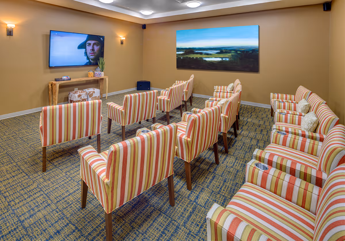 A cozy media room with multiple striped armchairs arranged in rows facing a wall-mounted flat screen TV. The room has beige walls, a patterned carpet, wall sconces for lighting, a wooden console table with decorative items, and a large landscape painting on the wall.