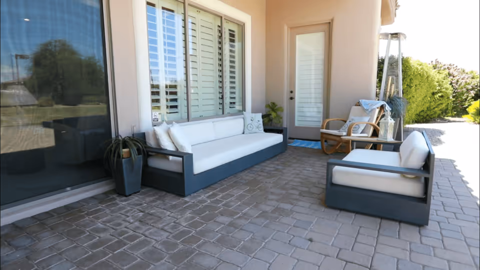 Outdoor patio area with cushioned seating including a long sofa, a single chair, and a wooden rocking chair. The patio has stone tile flooring and is adjacent to a building with a window and a door. There are plants and greenery visible in the background.