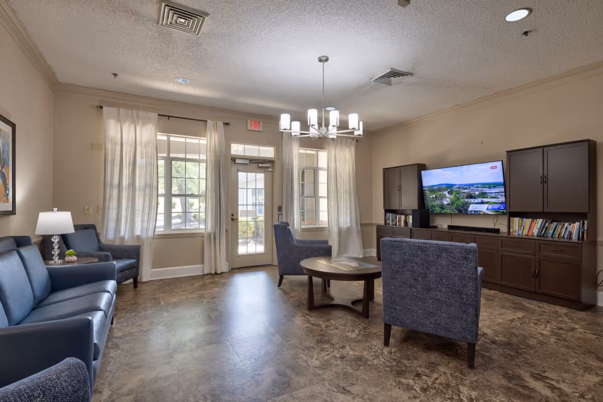 A senior living facility common area with blue upholstered chairs and a sofa arranged around a round wooden coffee table. A flat-screen TV is mounted on the wall above a wooden cabinet filled with books. Large windows with sheer curtains and a glass door allow natural light into the room. A modern chandelier hangs from the ceiling.