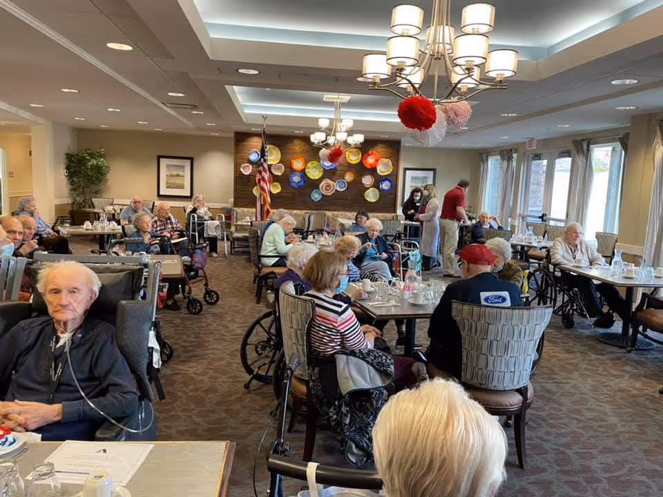 A dining room in a senior living facility with elderly residents seated at tables. Some residents are in wheelchairs, and a few staff members are attending to them. The room is well-lit with large windows, chandeliers, and colorful decorative plates on the far wall. An American flag is also visible in the background.