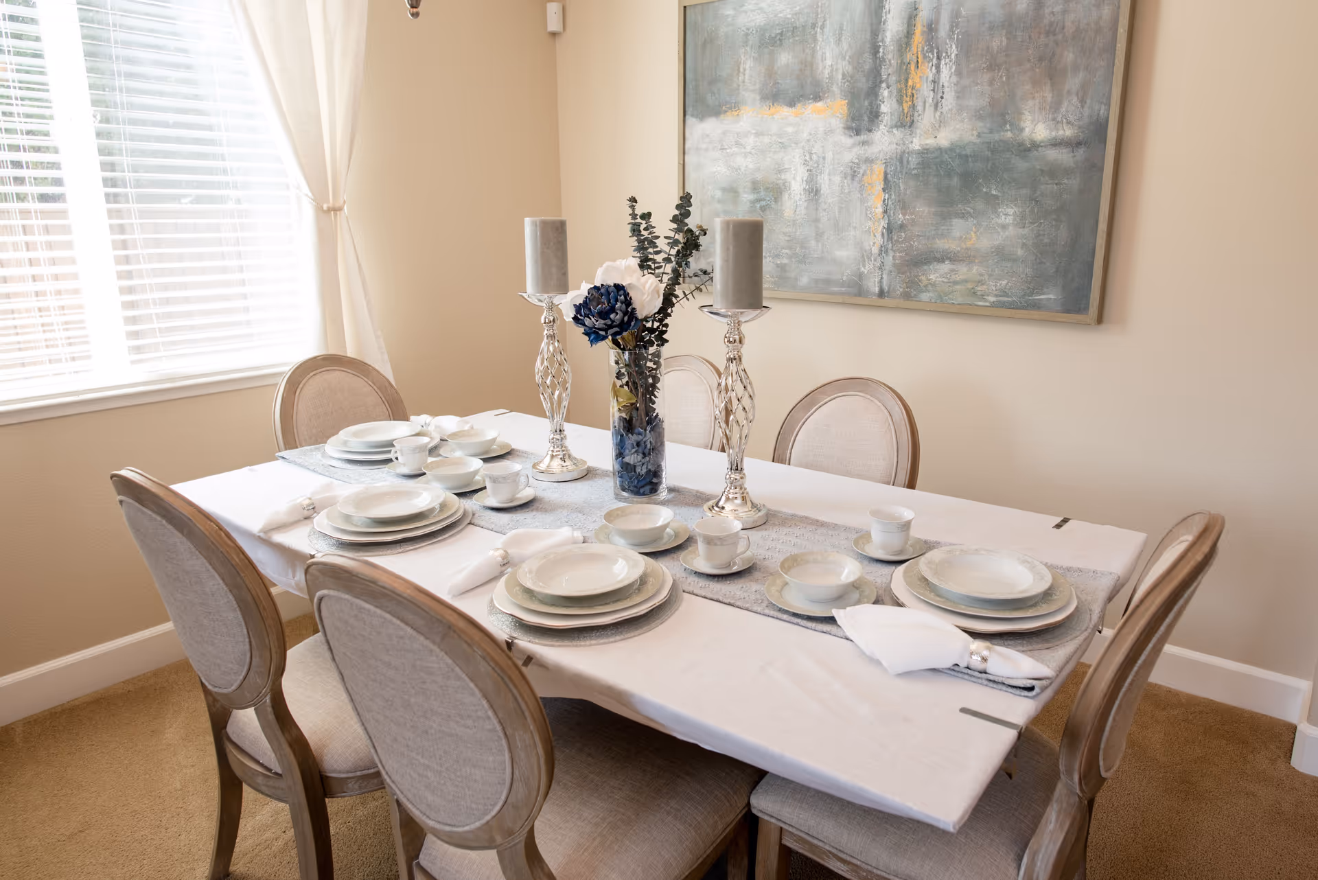 A dining room with a rectangular table set for six people. The table is covered with a white tablecloth and a gray runner, and is set with white plates, bowls, cups, and napkins with silver napkin rings. Two silver candle holders with gray candles and a vase with white and blue flowers are placed in the center. The room has beige walls, a large window with white blinds and curtains, and a large abstract painting on the wall.