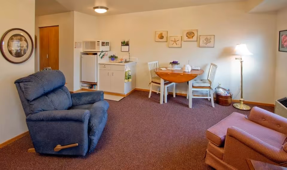 A cozy senior living room with a blue recliner chair and a pink armchair. There is a small dining table with two white chairs, a floor lamp, and a kitchenette area with a mini fridge, microwave, and sink. The walls are decorated with framed pictures and the carpet is brown.