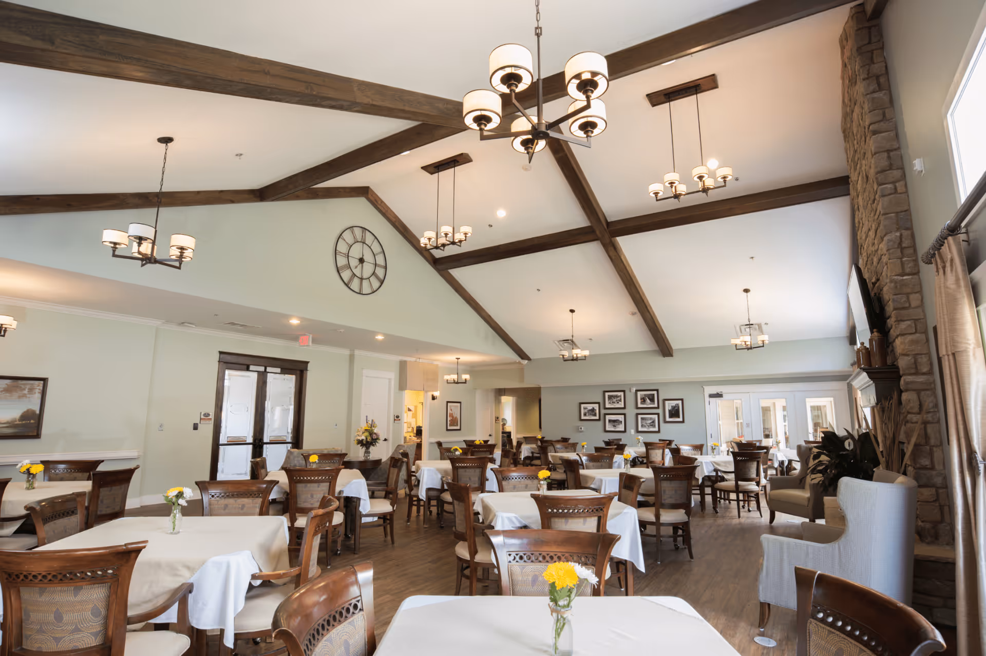 Spacious dining room with multiple set tables and chairs, vaulted ceiling with exposed wooden beams and chandeliers, and a stone fireplace on the right.