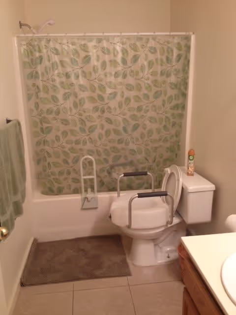 Bathroom with a bathtub and a shower curtain featuring a green leaf pattern. There is a toilet with safety rails on both sides and a raised toilet seat. A green towel hangs on the wall to the left, and a brown bath mat is placed on the tiled floor in front of the tub. A can of air freshener is on top of the toilet tank.