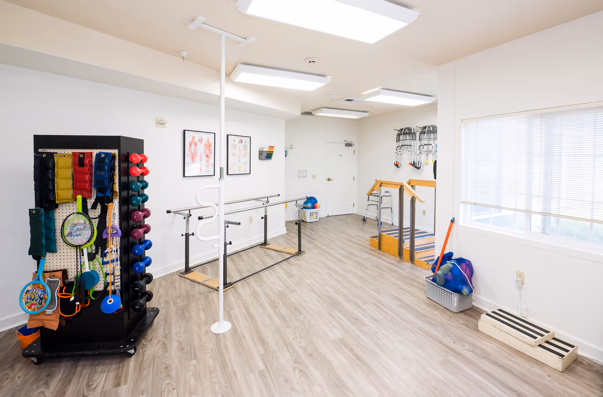 A bright physical therapy or rehabilitation room with light wood flooring and white walls. The room contains various exercise and therapy equipment including a rack with colorful weights and therapy tools, parallel bars for walking exercises, anatomical charts on the wall, a small set of stairs with handrails, and baskets with exercise balls and other equipment near a large window with blinds.