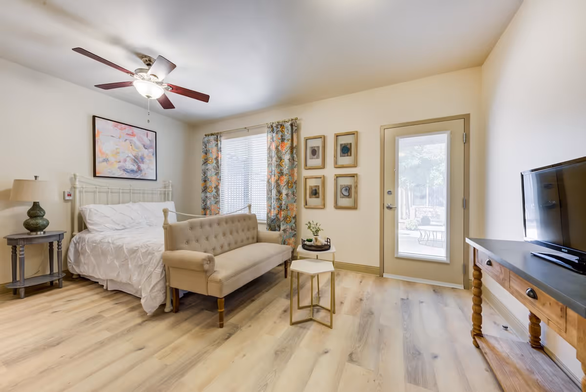 A bright and cozy bedroom featuring a white bed with a tufted beige bench at the foot, a ceiling fan with lights, a side table with a lamp, floral curtains on a window, four framed artworks on the wall, a glass door leading outside, and a wooden console table with a flat-screen TV.