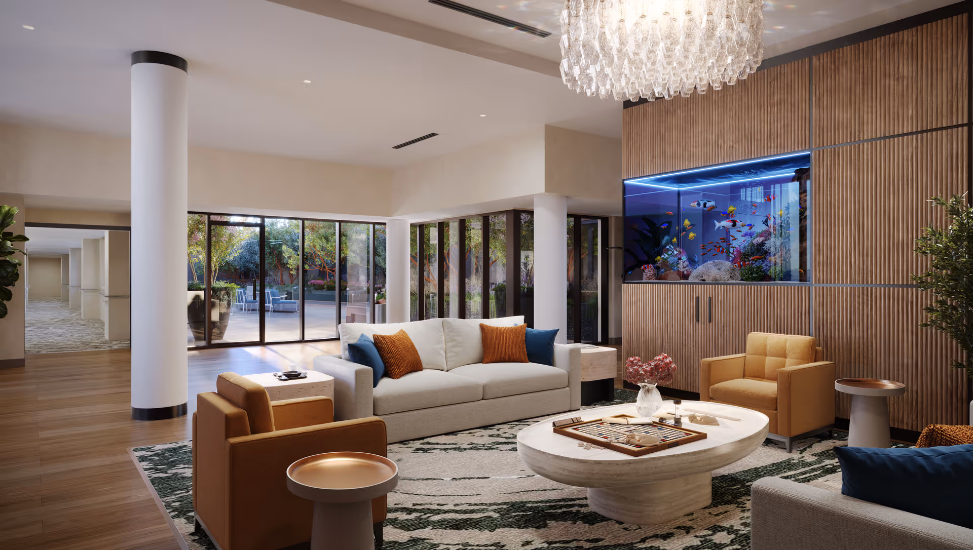 A modern living room with a white sofa and orange armchairs arranged around a white oval coffee table with a board game on it. There is a large built-in aquarium with colorful fish on the wooden paneled wall. Large windows and glass doors reveal an outdoor patio with seating and greenery. A crystal chandelier hangs from the ceiling.