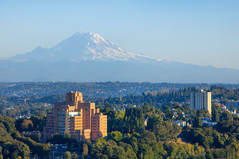 A brick multi-story building surrounded by trees with a snow-capped mountain dominating the distant skyline.