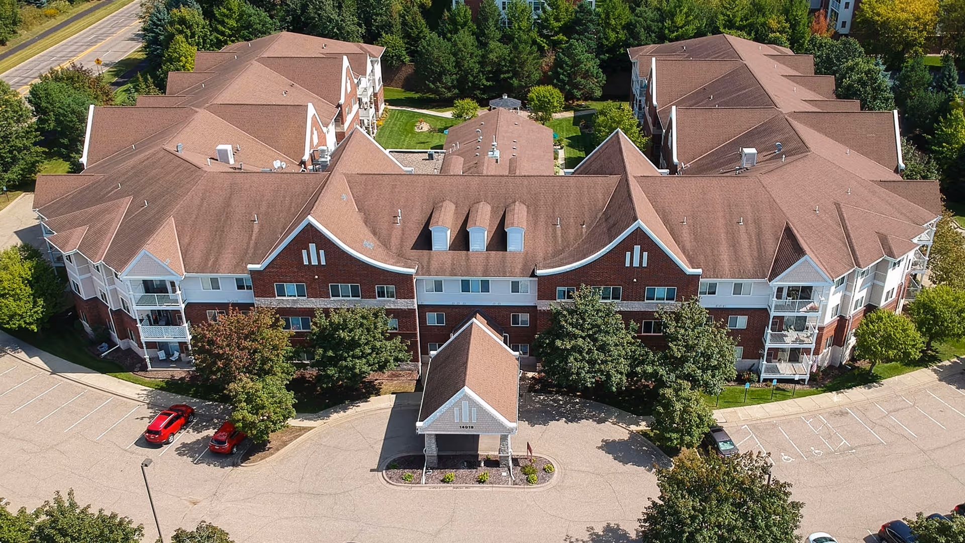 Aerial view of a large senior living facility building with a brown roof and red brick exterior. The building is surrounded by trees, parking spaces, and a driveway with a covered entrance. The facility is named The Timbers of Apple Valley.