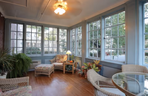 Sunlit sitting room with wicker chairs and floral cushions, a side table lamp, houseplants, and large multi-pane windows.