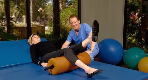 A woman lies on a padded exercise mat with a large cylindrical bolster under her knees while a man in blue scrubs assists her with a leg exercise. They are in a room with large windows showing trees and outdoor greenery.
