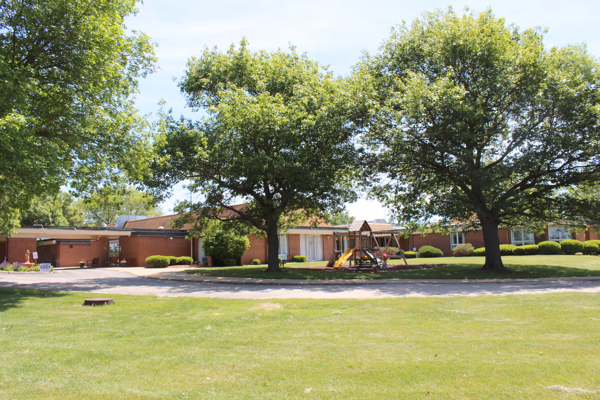 Exterior view of a single-story brick nursing home building with large green trees and a playground with a slide and swings in front. The area is surrounded by well-maintained grass and bushes under a clear sky.