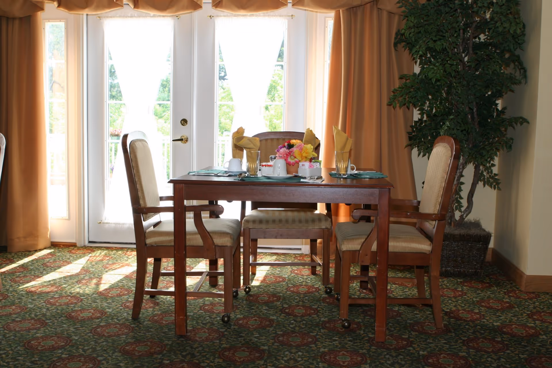 A dining area with a wooden table set for four, featuring beige cushioned chairs, folded yellow napkins in glasses, cups, and a small flower arrangement in the center. The room has patterned green and red carpet, large windows with sheer white curtains and beige drapes, and a tall potted plant in the corner.