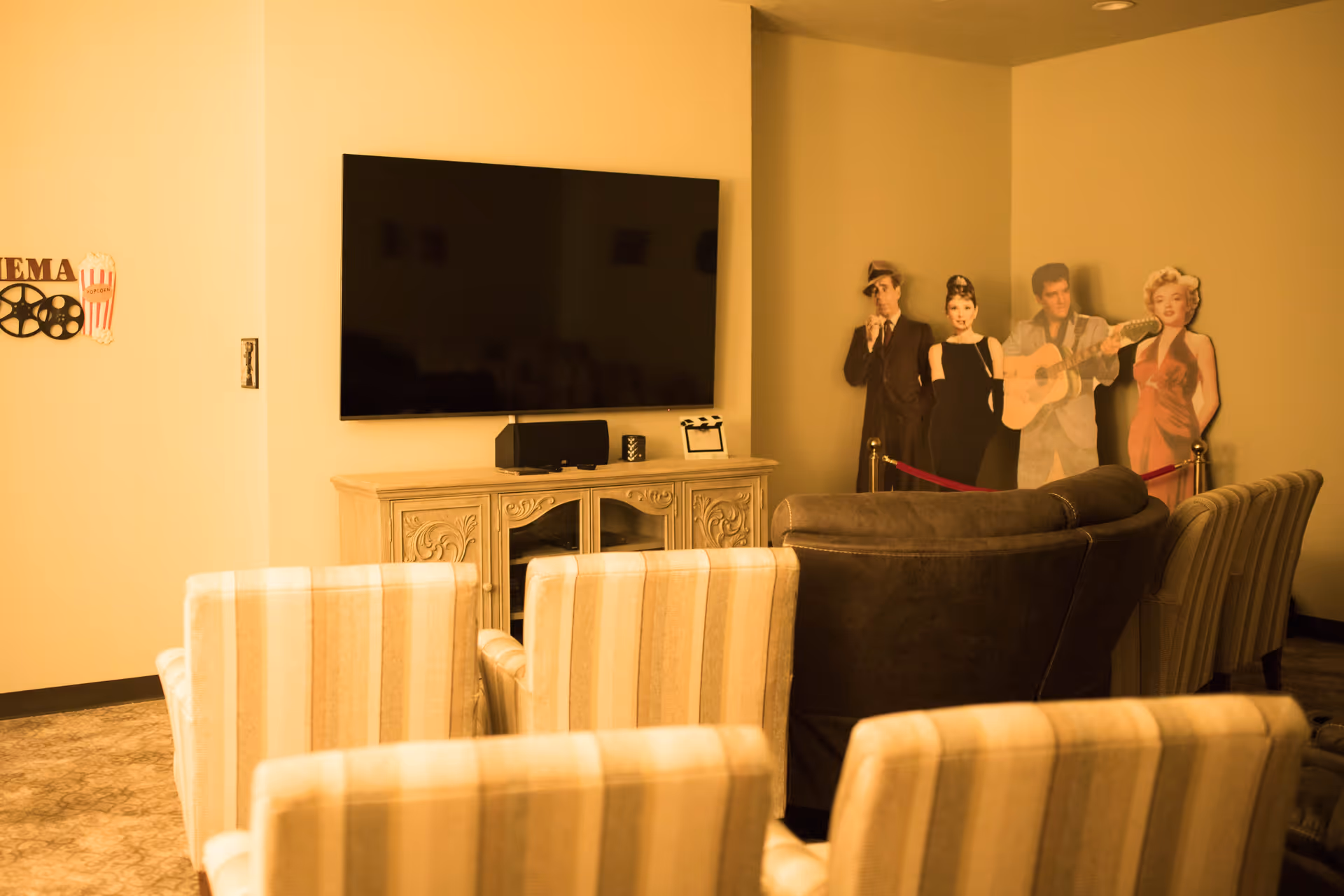 A cozy entertainment room with a large flat-screen TV mounted on a beige wall above a decorative wooden cabinet. In front of the TV are several striped upholstered chairs and a brown leather couch arranged for viewing. On the right side of the room, there are life-sized cardboard cutouts of classic movie stars roped off with a red velvet rope. A wall decoration with the word 'CINEMA' and a popcorn graphic is visible on the left wall.