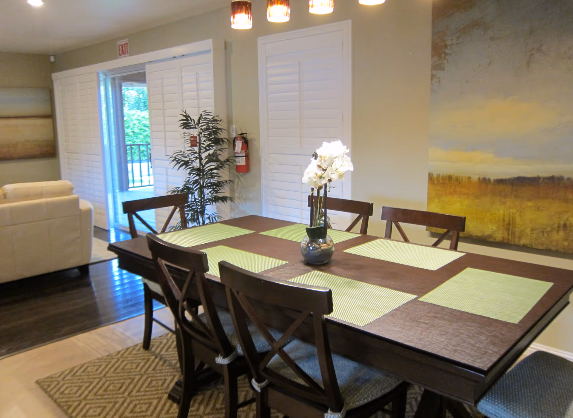 A dining area with a dark wooden table set with six chairs. The table has green placemats and a vase with white flowers in the center. In the background, there is a sliding glass door with white plantation shutters, a potted plant, a fire extinguisher on the wall, and a large painting with warm tones. Part of a beige sofa is visible to the left.