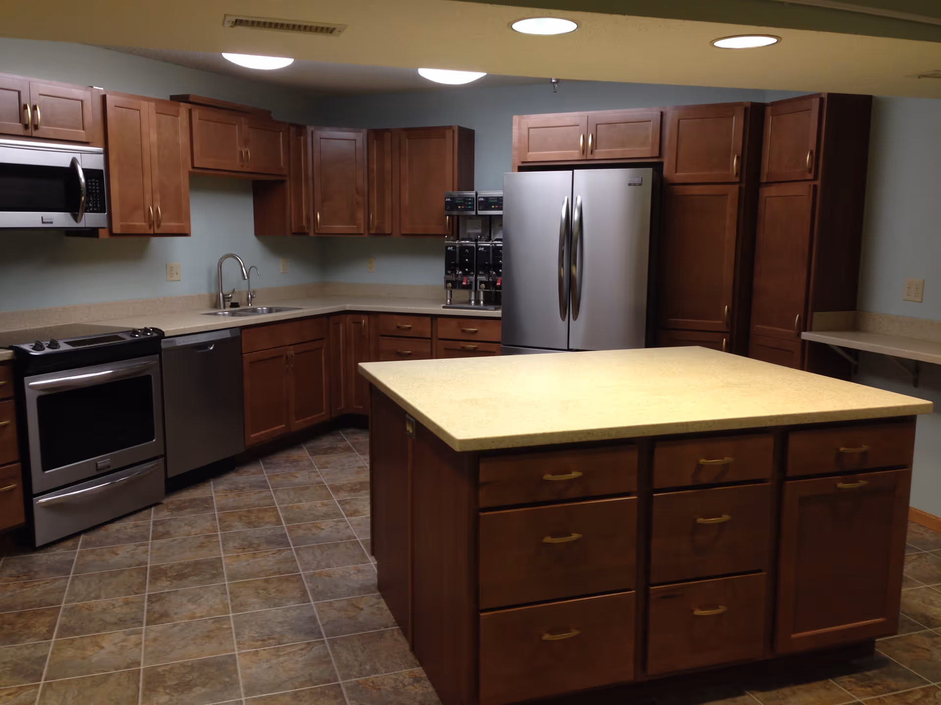 A spacious kitchen with wooden cabinets, a large central island with drawers, a stainless steel refrigerator, oven, microwave, dishwasher, and a coffee machine. The floor is tiled, and the walls are painted light blue.