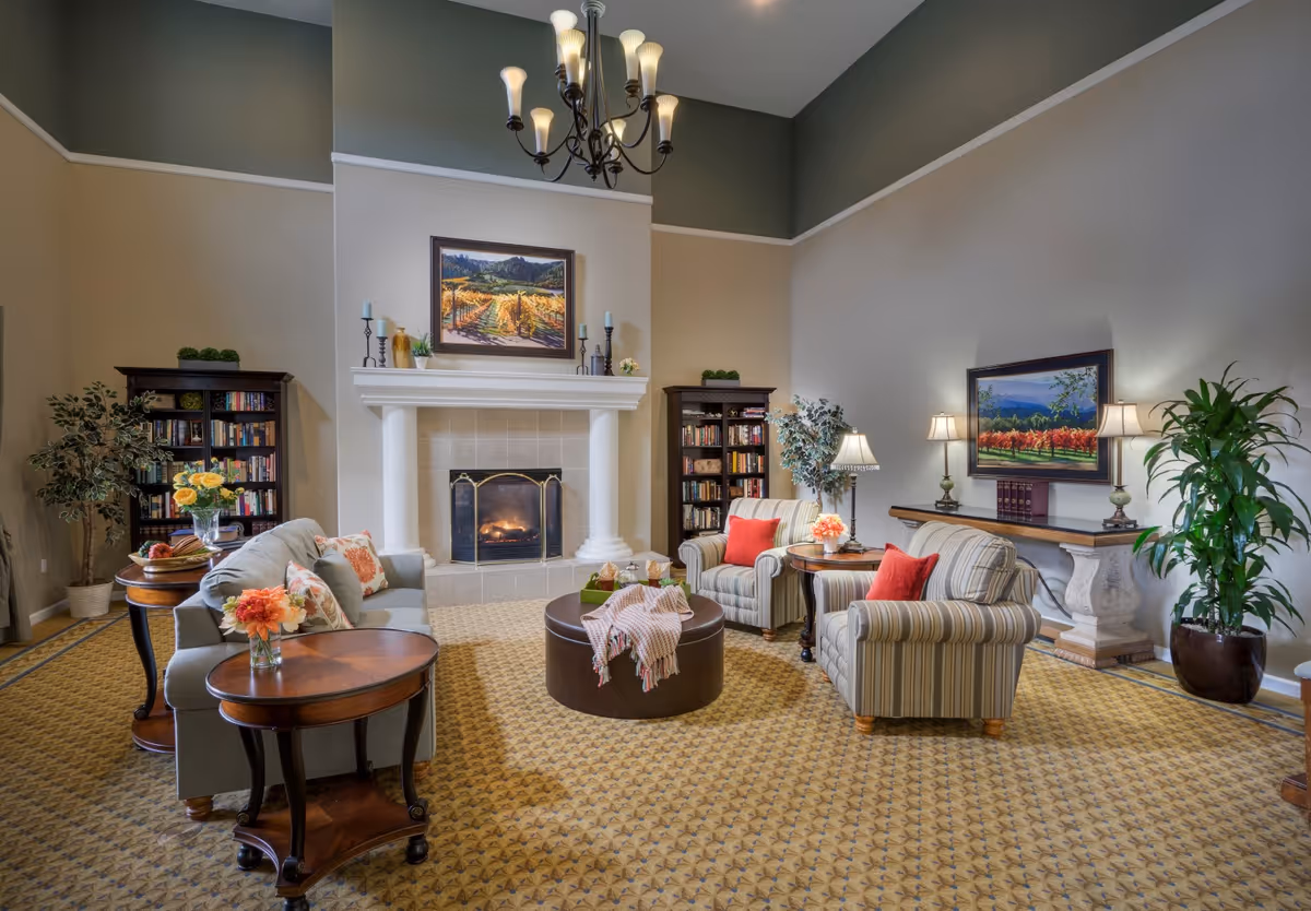 A cozy living room with a lit fireplace centered between two dark wood bookshelves filled with books. The room features a light blue sofa with floral pillows, two striped armchairs with red cushions, wooden side tables with flower arrangements, a round ottoman with a throw blanket, and a chandelier hanging from the ceiling. The walls are painted in two tones with framed landscape paintings and several lamps providing warm lighting.