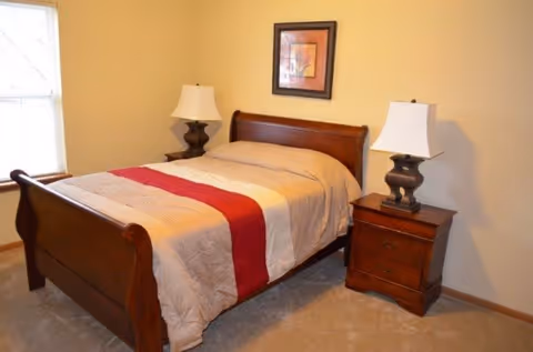 A bedroom with a wooden bed frame featuring a beige bedspread with a red stripe down the middle. There are two matching wooden nightstands on either side of the bed, each with a lamp. A framed picture hangs on the wall above the bed, and a window with blinds is visible on the left side.