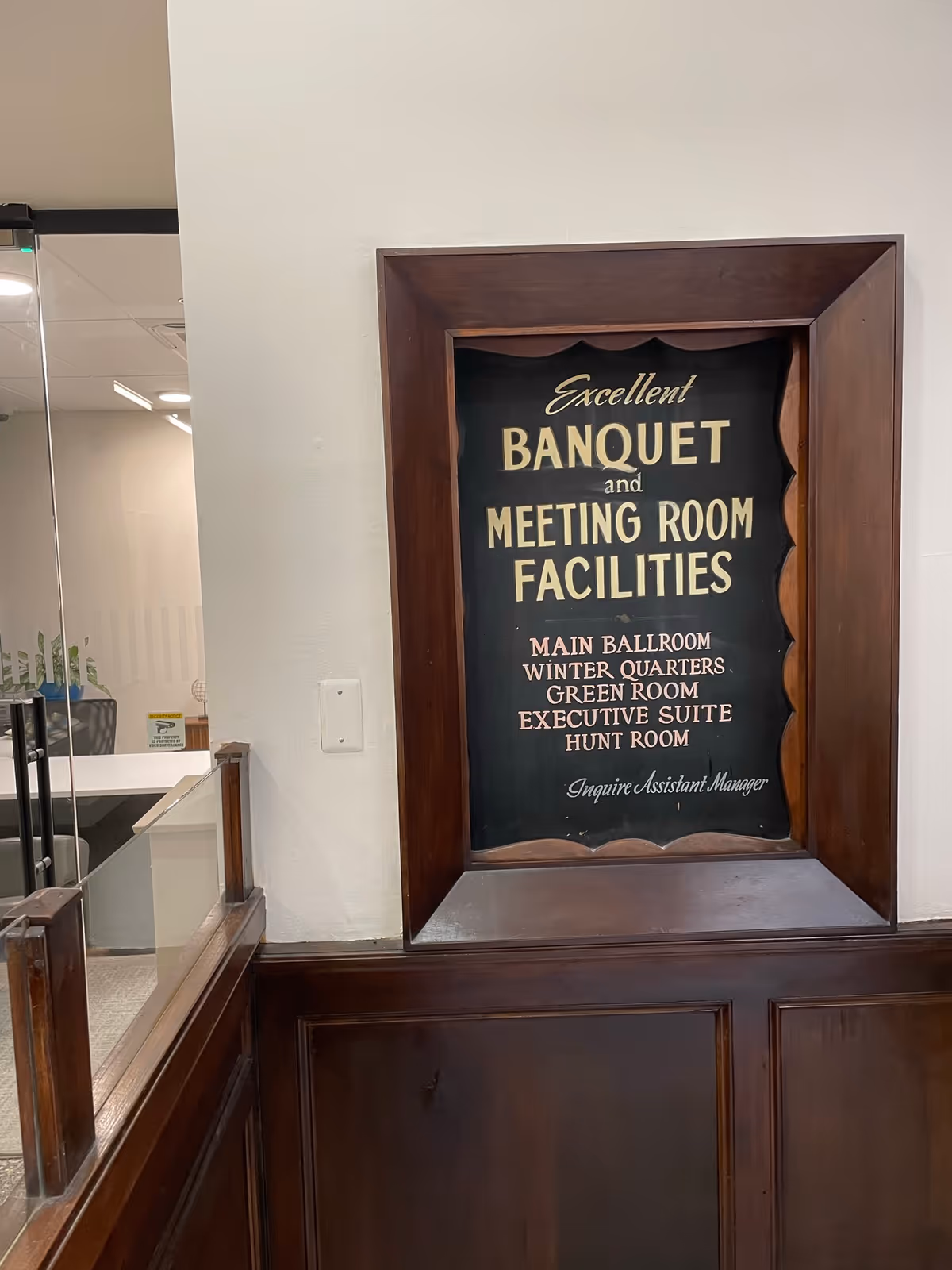A framed sign on a wall inside a building that reads 'Excellent Banquet and Meeting Room Facilities' with a list of rooms including Main Ballroom, Winter Quarters, Green Room, Executive Suite, and Hunt Room. The sign is mounted above dark wood paneling and there is a glass partition to the left.