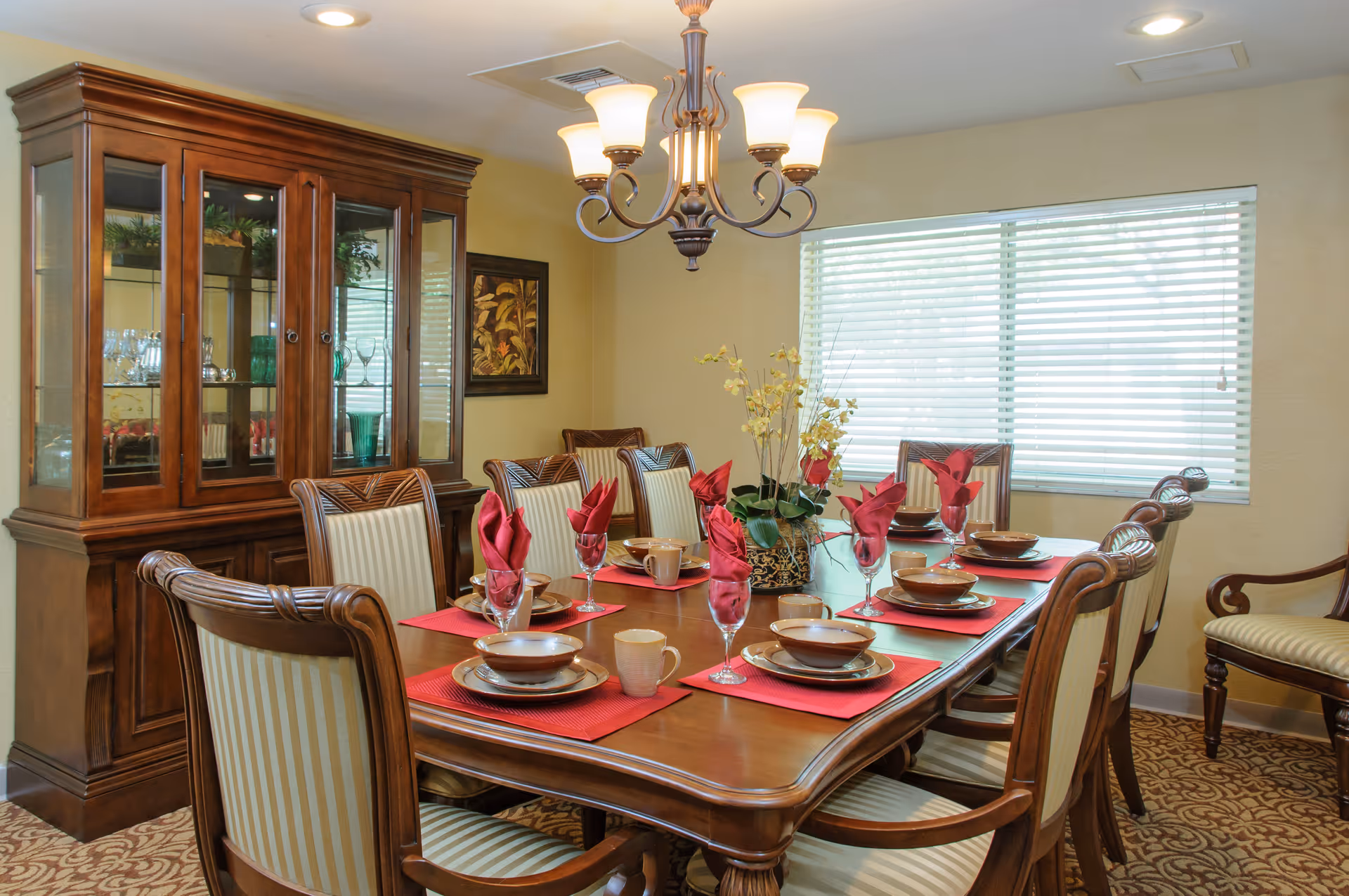 A formal dining room with a large wooden dining table set for eight people. The table has red placemats, brown dishes, cups, and wine glasses with red folded napkins. A floral centerpiece is in the middle of the table. The room features a wooden china cabinet with glass doors displaying glassware, a window with blinds, a chandelier overhead, and patterned carpet flooring.