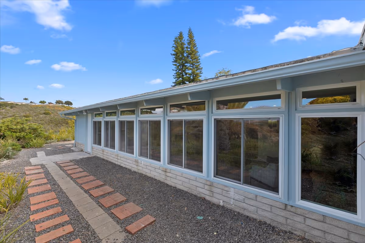 Side exterior of a single-story light-blue building with a row of large windows and a brick-and-gravel walkway