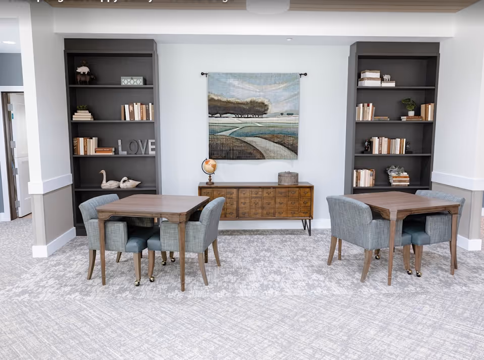 Bright common room with two wooden tables and upholstered chairs flanked by built-in bookshelves and a landscape wall hanging.