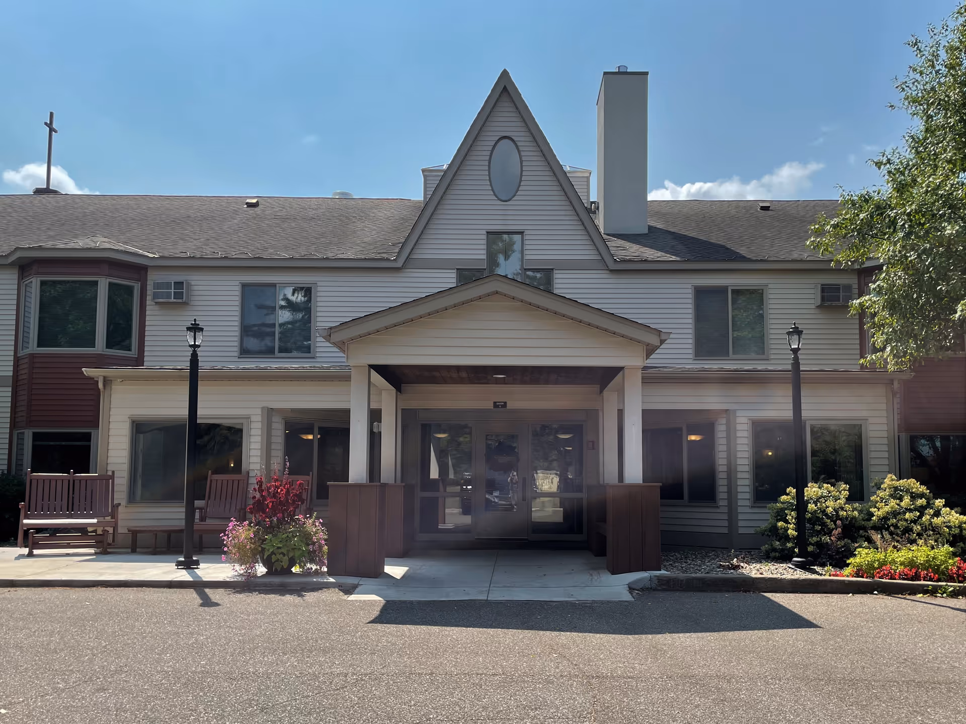 Front exterior view of Benedict Village | An Ecumen Living Space building with a covered entrance, two lamp posts, benches, and landscaping including flowers and bushes under a clear blue sky.