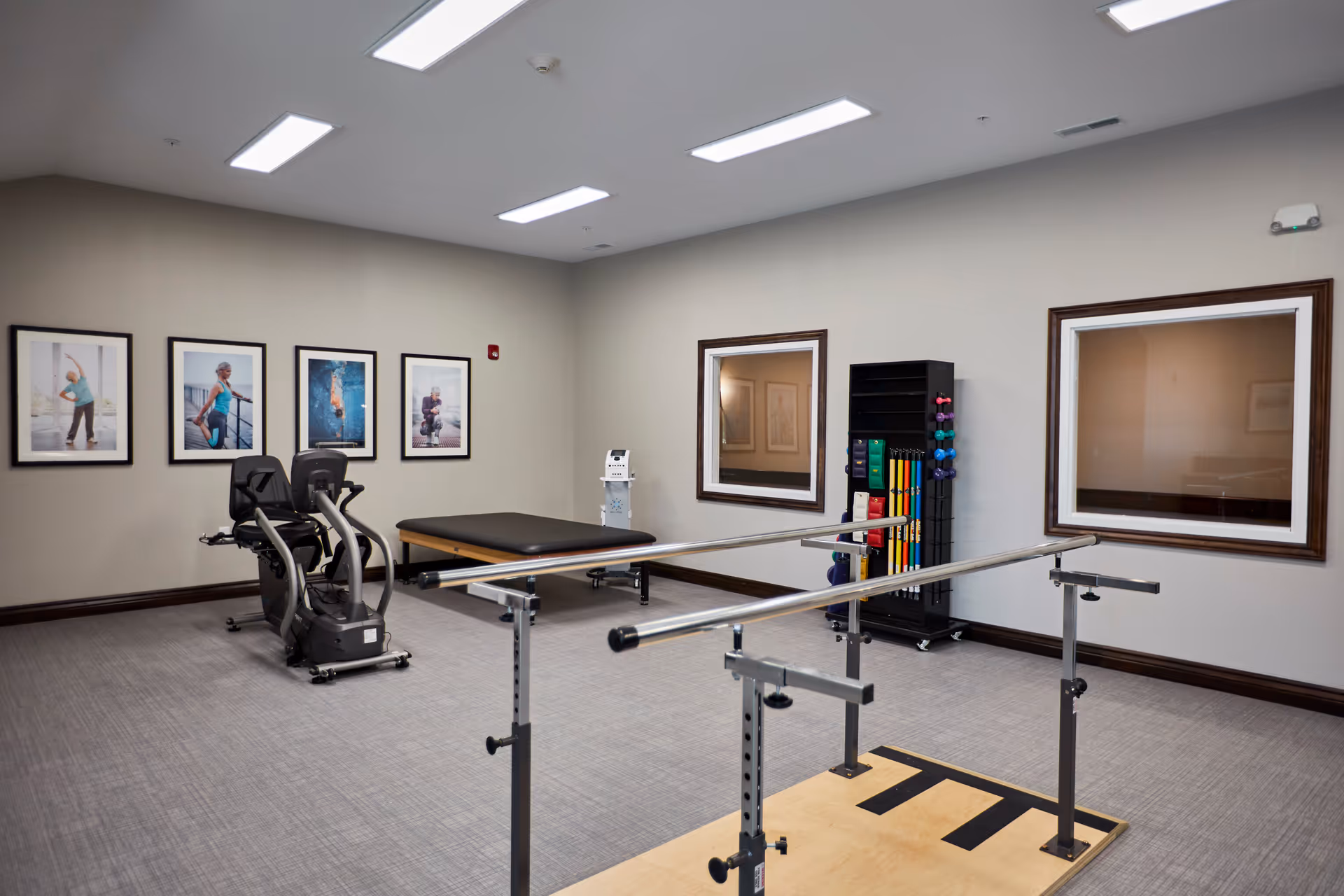 A senior living facility exercise room with parallel bars for walking support, a recumbent exercise bike, a padded therapy table, and a rack holding various exercise equipment such as dumbbells and resistance bands. The walls are decorated with framed photos of seniors exercising.