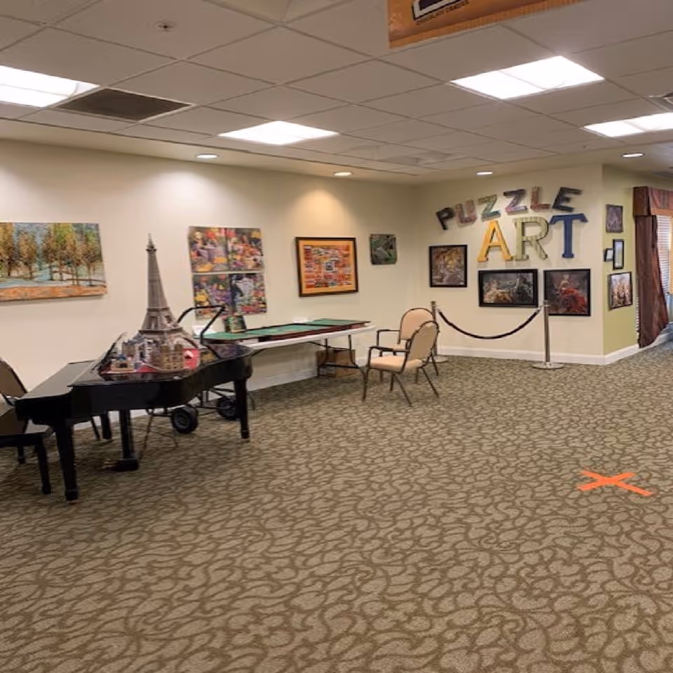 Interior room with a patterned carpet and beige walls decorated with various framed pictures and the words 'PUZZLE ART' on the wall. There is a black grand piano with a model of the Eiffel Tower on top, a table with puzzle pieces, and two chairs. The room is well-lit with ceiling lights.