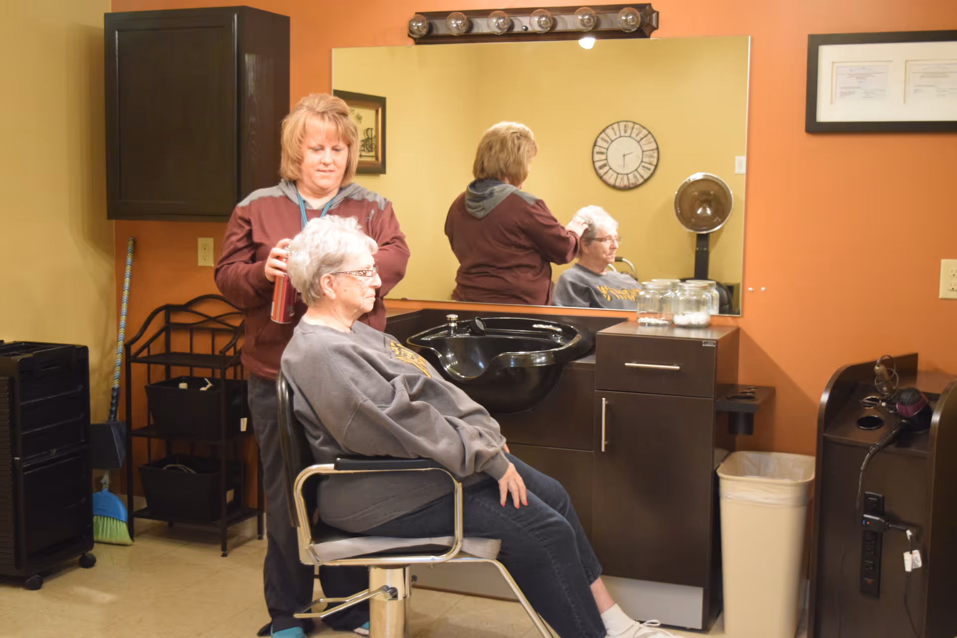 A senior woman with gray hair sits in a salon chair while a hairstylist stands behind her, holding a can of hair spray. They are in a hair salon area with a large mirror, a black sink, and cabinets. The reflection of both women is visible in the mirror. The room has warm-colored walls and various salon equipment.