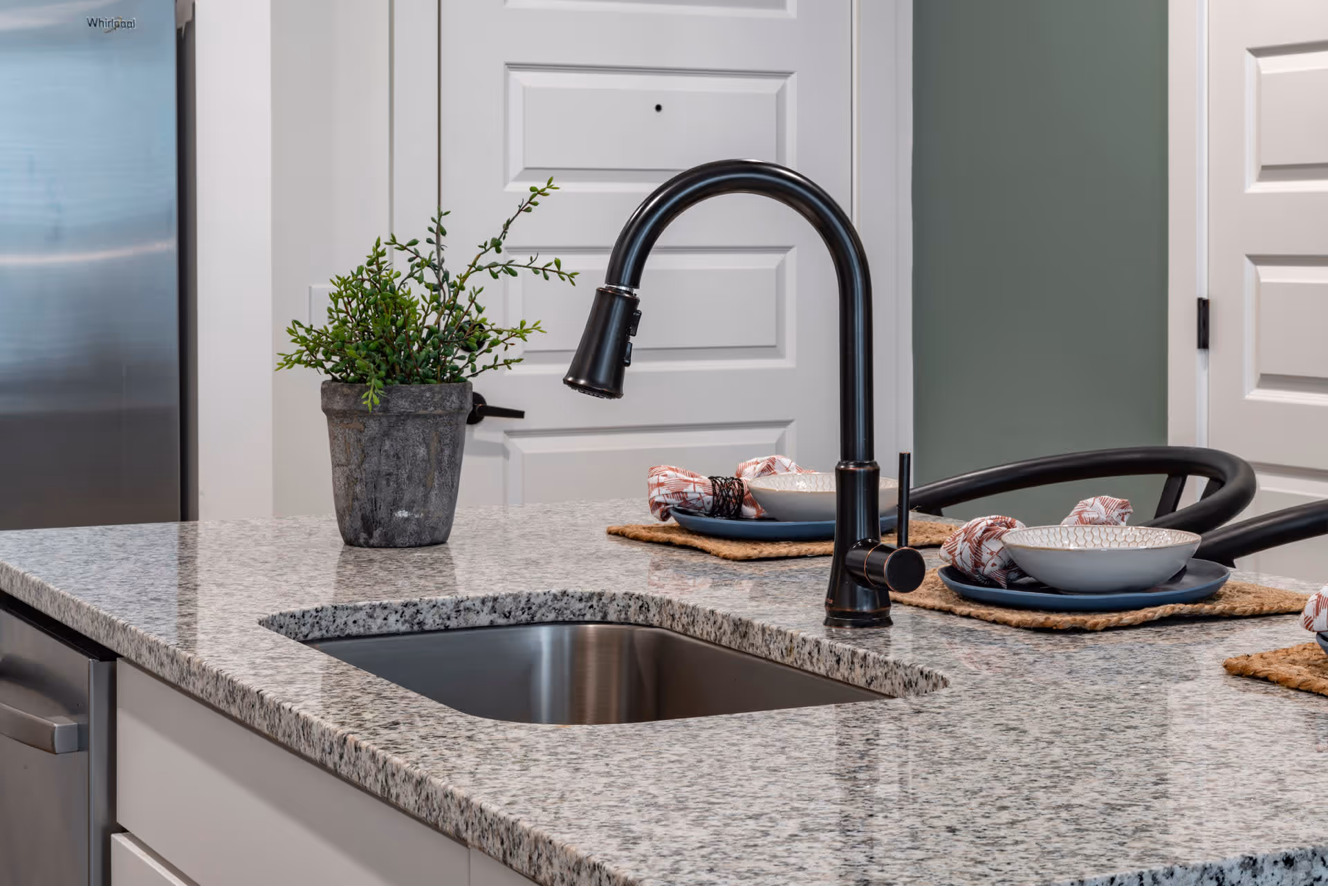Modern kitchen countertop with a stainless steel sink and black faucet. On the granite countertop, there is a small potted plant and two place settings with bowls, plates, and napkins on woven placemats. In the background, there is a stainless steel refrigerator and white paneled doors.
