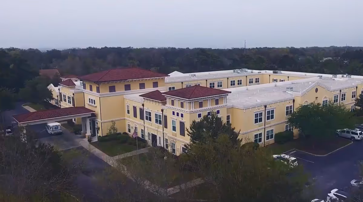 Aerial view of a large, two-story yellow building with a red-tiled roof surrounded by trees and parking areas, identified as The Beacon at Gulf Breeze.