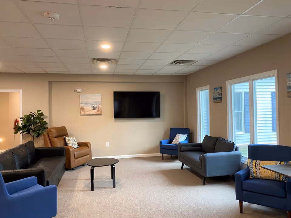 A cozy living room with beige walls and carpeted floor featuring a black leather sofa, a brown recliner, two blue armchairs, and a gray loveseat. A small round black coffee table is in the center. A flat-screen TV is mounted on the wall above a beach-themed painting. Large windows on the right side let in natural light.