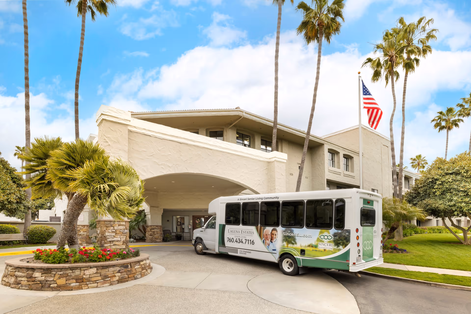 Exterior view of Laguna Estates Senior Living facility with a shuttle bus parked in front. The building is beige with a covered entrance, surrounded by palm trees and landscaped greenery. An American flag is flying on a flagpole near the entrance.