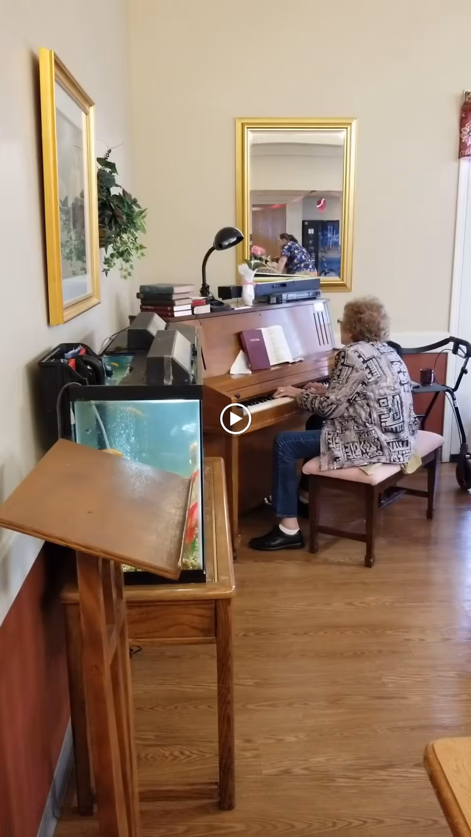 An elderly woman sits at an upright piano in a communal interior space with a fish tank, mirror, and a walker nearby.