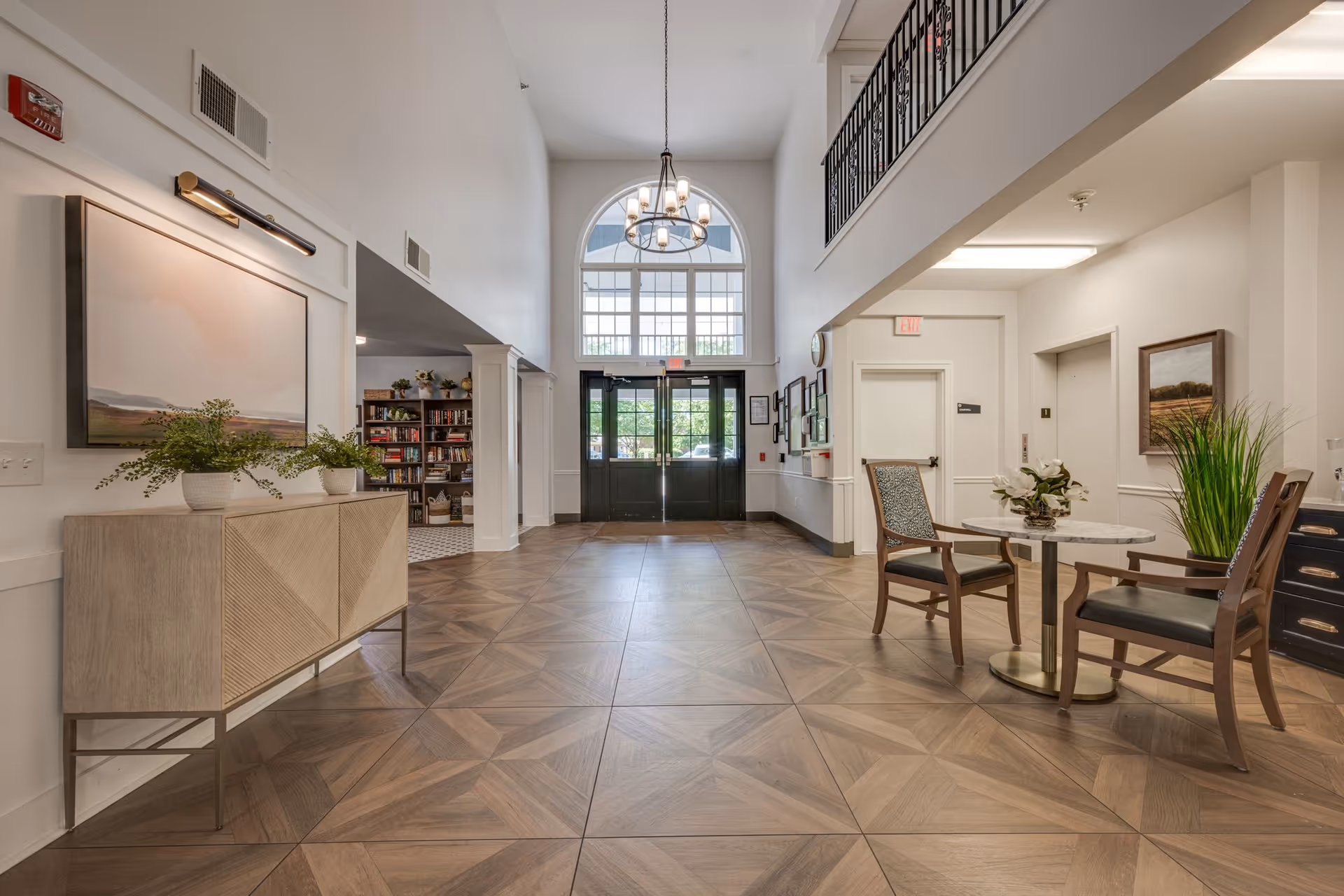 Spacious and well-lit interior lobby area of an assisted living facility with a high ceiling, large arched window above double glass doors, wooden patterned floor tiles, a small round table with two chairs, plants, artwork on the walls, and a bookshelf visible in an adjacent room.