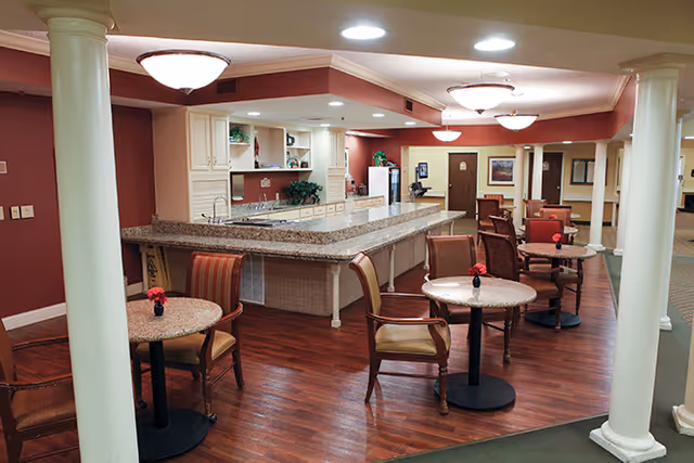 Interior view of a senior living facility common area with a large granite countertop kitchen island, several small round tables with chairs, red walls, white columns, and ceiling lights. The space has wooden flooring and decorative plants on the kitchen shelves.